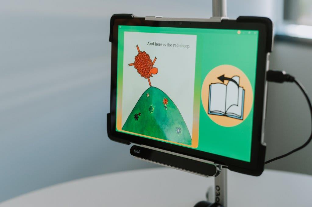 Mounted iPad or table showing AAC program with sheep doing handstand and book on screen